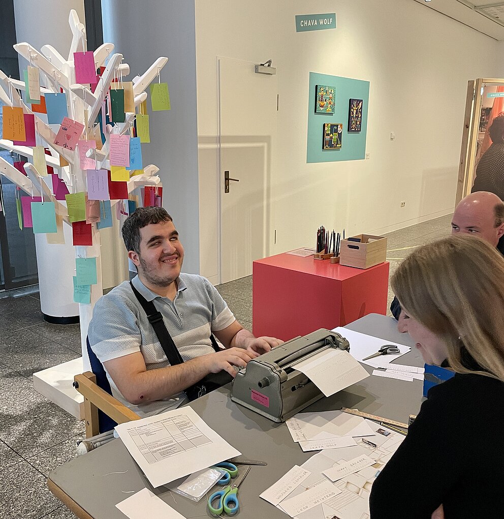 Personen sitzen an einem Tisch mit viel Papier. Eine Person schreib an einer Schreibmaschiene. 