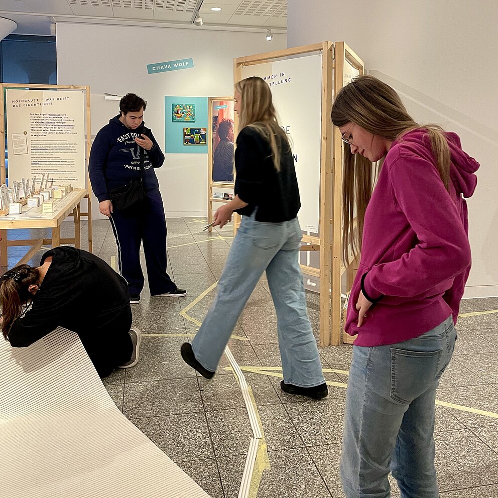 Verschiedene Personen stehen und laufen um menschengroße Holztafeln. Auf dem Boden sind wenige weiße Streifen.